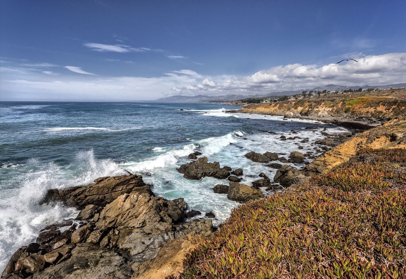 A scenic view of a rocky coastline with waves crashing against the shore under a blue sky with clouds.
